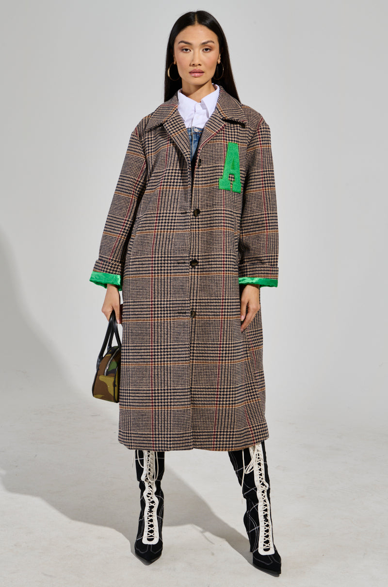 A woman wearing the TAKE IT EASY RELAXED FIT TRENCH, crafted from a wool-blend and showcasing a long plaid design with green accents, seamlessly pairs it with a white shirt and patterned boots. She holds a camouflage-patterned bag while standing against a plain background, looking directly at the camera.