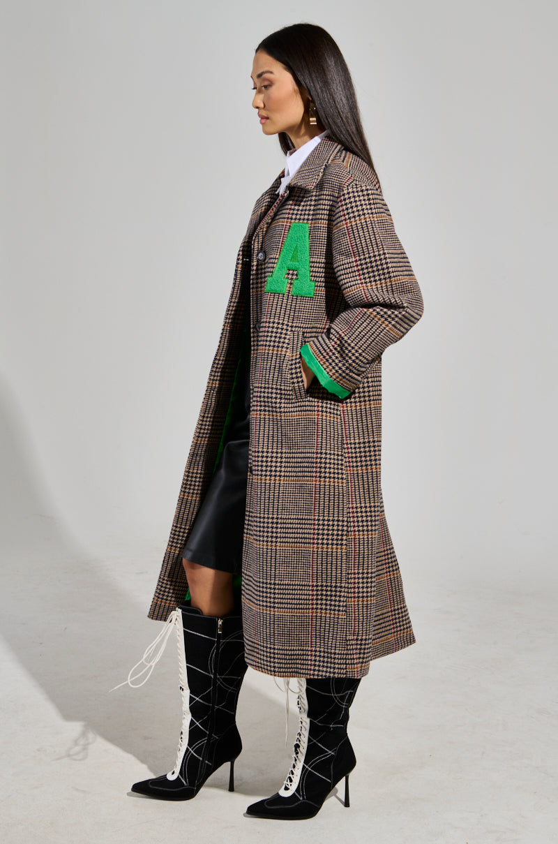 A woman is dressed in the TAKE IT EASY RELAXED FIT TRENCH, a long, checkered coat featuring a relaxed fit and a green letter "A" on the front. Her ensemble is completed with a white shirt, black skirt, and knee-high black lace-up boots with white laces. She stands in profile view against a plain background, exuding effortless style.