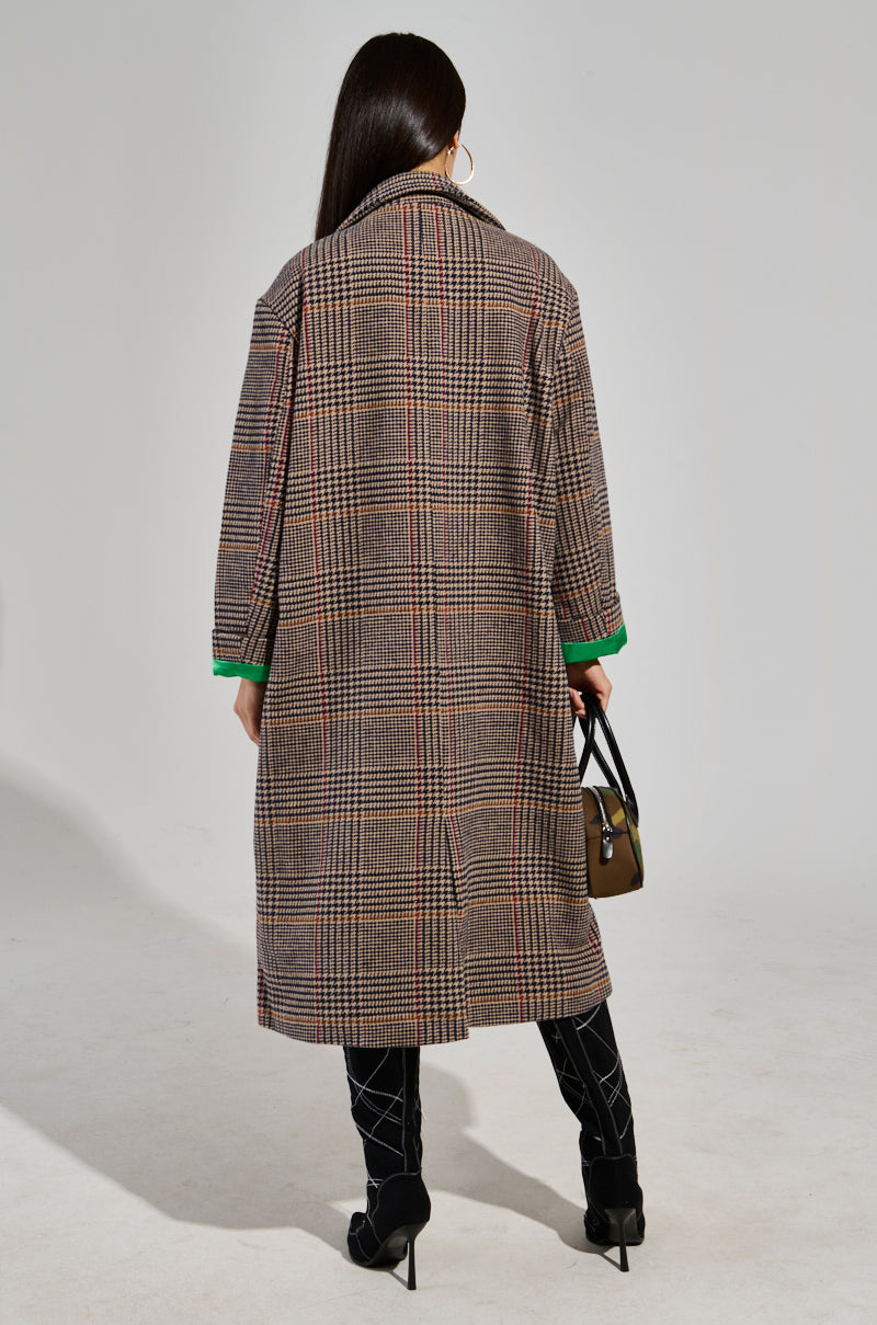 A person stands with their back to the camera, dressed in the TAKE IT EASY RELAXED FIT TRENCH, a plaid coat crafted from a wool-blend fabric with green cuffs. They carry a small handbag and wear black pants featuring a geometric pattern paired with high-heeled shoes.