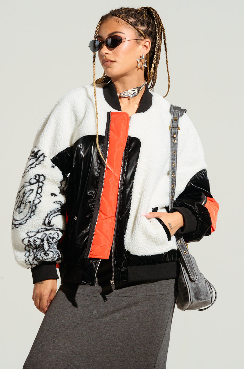 Wearing a fashionable urban chic outfit, the individual effortlessly combines white, black, and orange hues using graphic designs, featuring a pair of sunglasses and the LENNIE MIXED MEDIA BOMBER—a stylish oversized faux sherpa jacket. Posing against a neutral backdrop with a gray shoulder bag and skirt as accessories, they achieve the ultimate modern look.