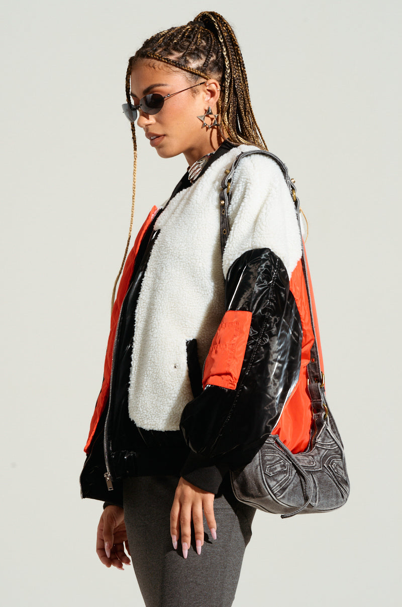 A person showcases an urban chic ensemble, wearing LENNIE MIXED MEDIA BOMBER—a stylish two-toned jacket in white and red with black sleeves—and a pair of sunglasses. They carry a patterned shoulder bag and flaunt long braided hair adorned with star-shaped earrings. The plain backdrop emphasizes their striking style.