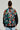 Someone with their back turned is wearing the TALK TO ME NICE BOMBER, a jacket featuring graffiti-inspired designs and vibrant colors. The individual has their hair tied up and is also sporting large hoop earrings. Holding a bag in their left hand, they epitomize urban chic outerwear.