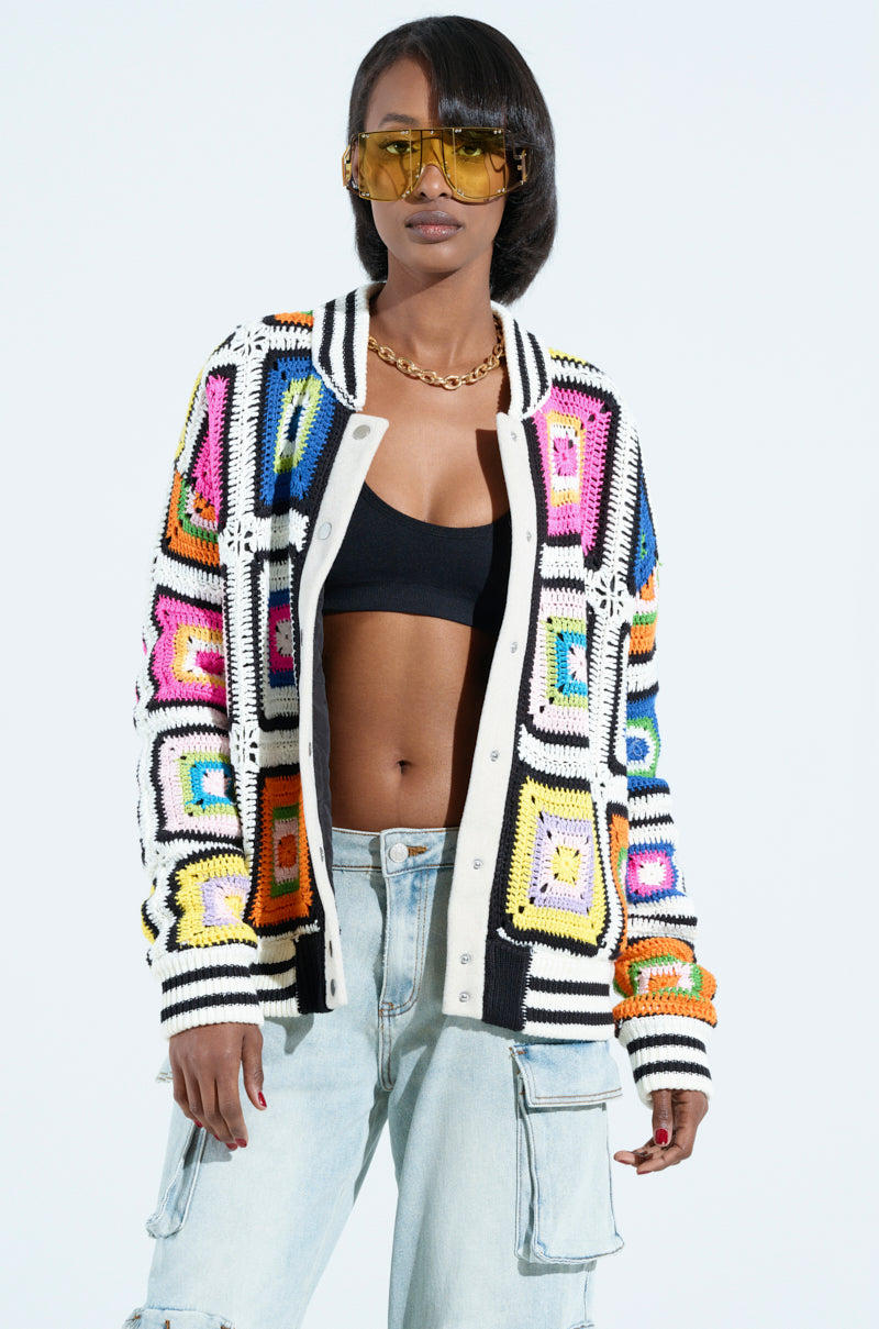 Against a plain backdrop, an individual wears the ALHAMBRA CROCHET BOMBER, adorned with colorful granny squares, complemented by a black top, light blue jeans, and yellow-tinted sunglasses.