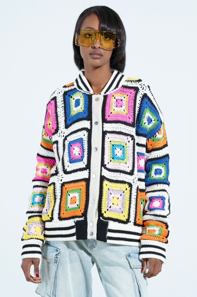 Someone in oversized square sunglasses wears the ALHAMBRA CROCHET BOMBER, adorned with colorful geometric granny square patterns. This vibrant cardigan with pink, orange, blue, and green hues is paired with light blue jeans against a plain background.