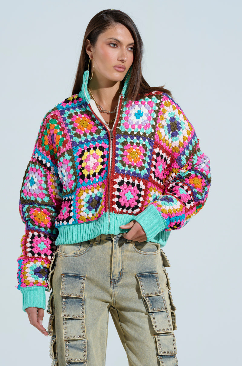 Someone is wearing the AZURRA CROCHET BOMBER, featuring a colorful, sherpa-lined crochet design. They stand against a plain background with long hair and wear light denim cargo pants with multiple pockets and zipper details.