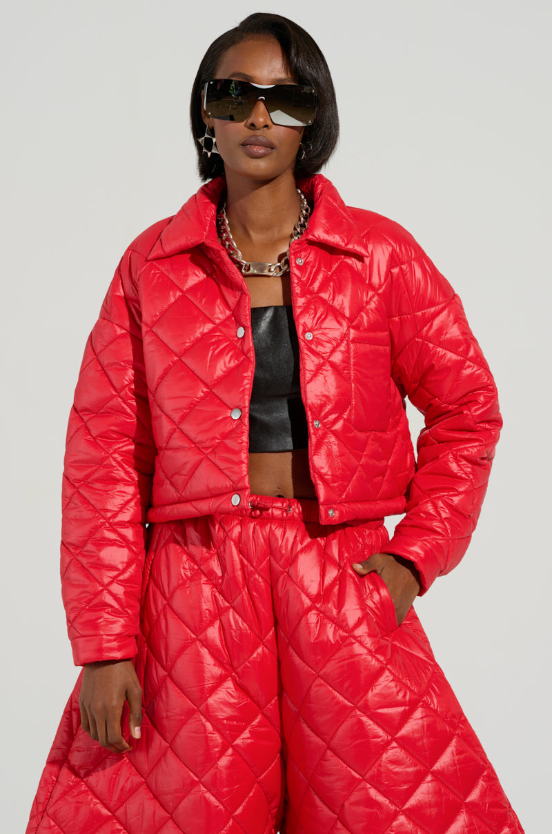 Dressed in the VIVI Cropped Puffer Bomber in Red, an individual showcases a striking look with matching wide-leg pants, complemented by a black top and sunglasses. The outfit is accessorized with a chunky chain necklace and set against a simple light gray background.
