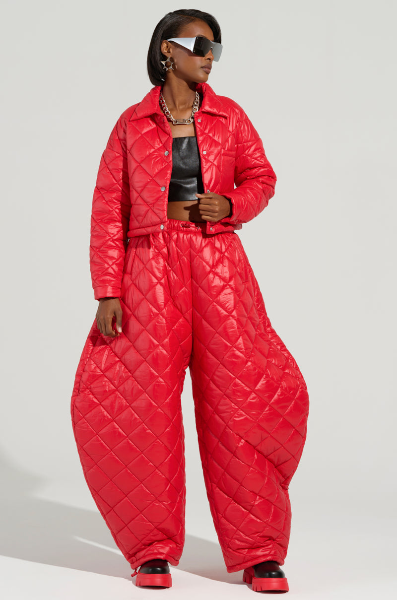 A person is dressed in an AKIRA Label Vivi outfit featuring the VIVI CROPPED PUFFER BOMBER IN RED and oversized quilted red pants. A black crop top peeks underneath, paired with sunglasses and red shoes. Their confident pose is accentuated by a neutral background.