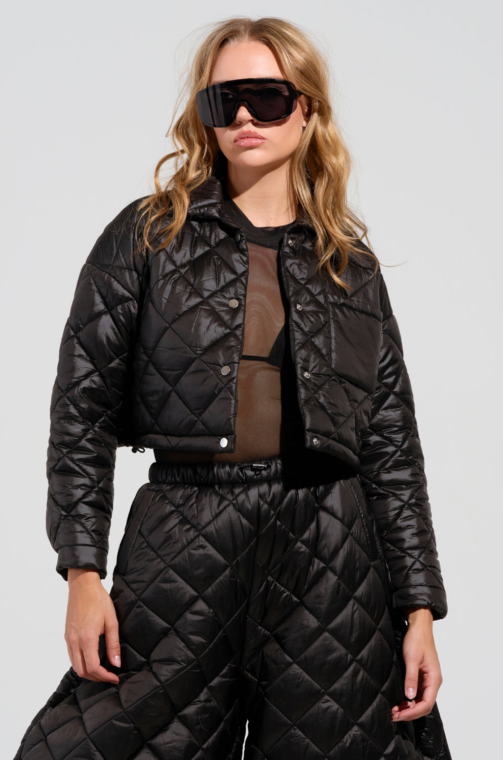 A woman wearing oversized black sunglasses and a VIVI Cropped Puffer Bomber in Black, paired with a mesh top and matching quilted pants. She has wavy blonde hair and is standing against a plain background.