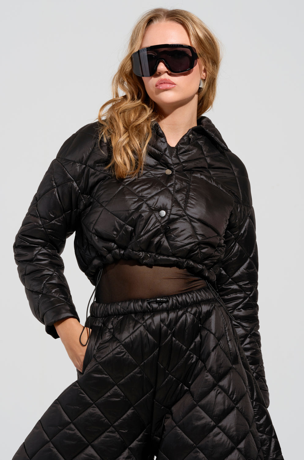 A person wearing the VIVI Cropped Puffer Bomber in black, paired with matching pants and large black sunglasses. They have long, wavy hair and are standing against a plain background, showcasing a stylish, modern look with its sleek inner lining.