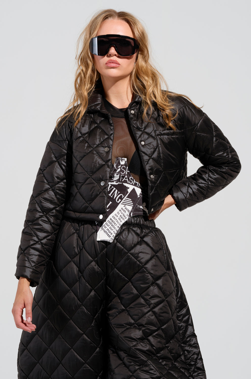 A person wearing oversized sunglasses and a VIVI Cropped Puffer Bomber in black poses confidently. Their outfit includes a graphic black top and matching quilted windbreaker pants, set against a plain background.