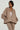 A person wearing a SOFIA FAUX SUEDE BOMBER in taupe along with matching pants stands against a neutral background. The oversized jacket, featuring large pockets, perfectly complements their shoulder-length hair and gold hoop earrings, finished with a chunky necklace for that cozy outerwear look.