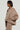 Wearing the oversized tan SOFIA FAUX SUEDE BOMBER from the AKIRA Label paired with matching pants, a person exudes effortless style. With shoulder-length hair and large gold hoop earrings, they accessorize flawlessly. The bomber is zipped partially to reveal a white top underneath, making it perfect cozy outerwear for any occasion.