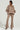 A person stands confidently in cozy outerwear, wearing a taupe SOFIA FAUX SUEDE BOMBER jacket and matching flared pants. They complete the ensemble with a white top, gold necklace, and textured beige ankle boots against a plain light gray background.