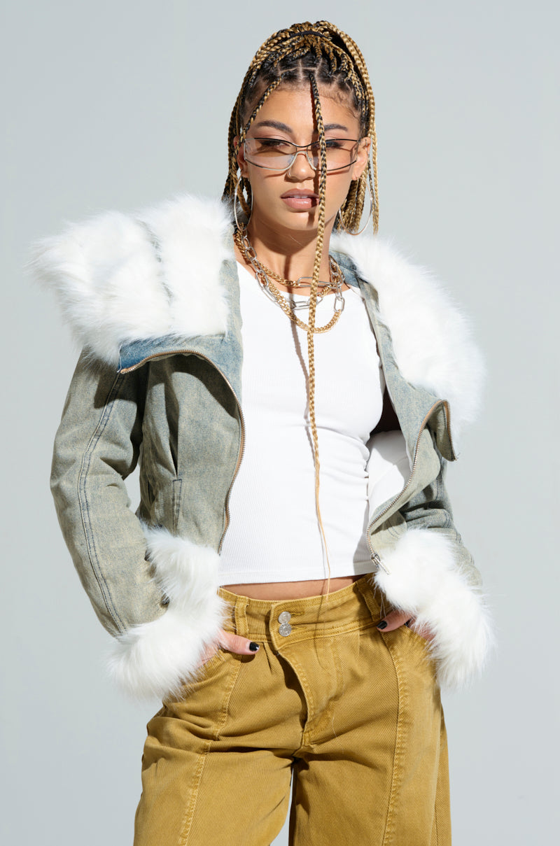 Dressed in the BELLA FUR TRIM DENIM MOTO jacket with its distinctive white fur trim, a person with braided hair and sunglasses exudes confidence. They pair the jacket elegantly with a white sleeveless top and mustard-colored pants, standing against a light gray background with one hand casually placed in their pocket.
