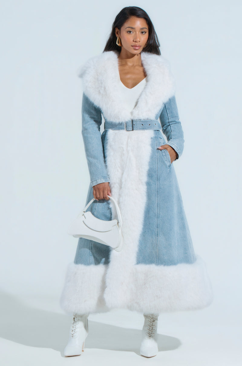 A person poses against a light blue background wearing the VIVIAN FUR TRIM COAT IN DENIM, featuring a faux fur trim and adjustable waist belt. They pair it with white high-heeled boots, hold a small white handbag, and have long hair.