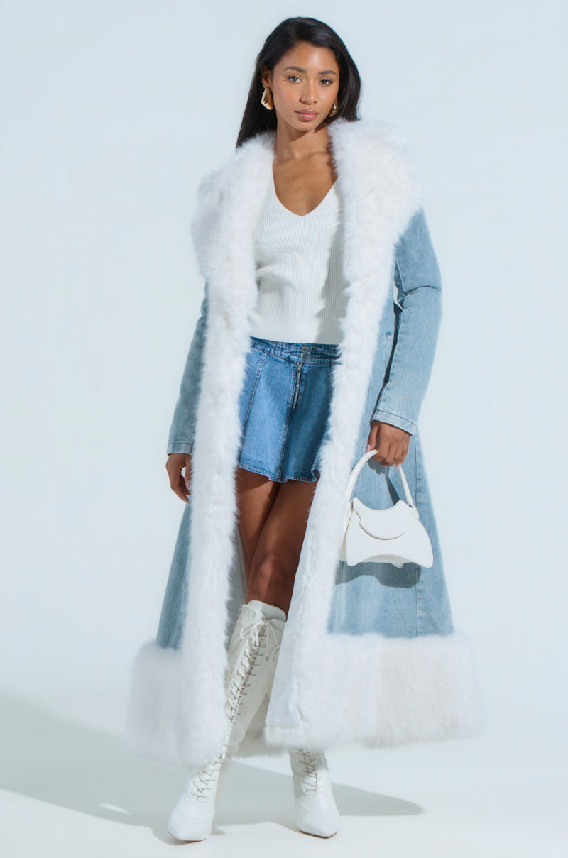 A woman models the "VIVIAN FUR TRIM COAT IN DENIM," featuring a long light blue design with an adjustable belt and white faux fur trim. She pairs it with a white top, denim skirt, knee-high white lace-up boots, and carries a small white handbag against a plain background.