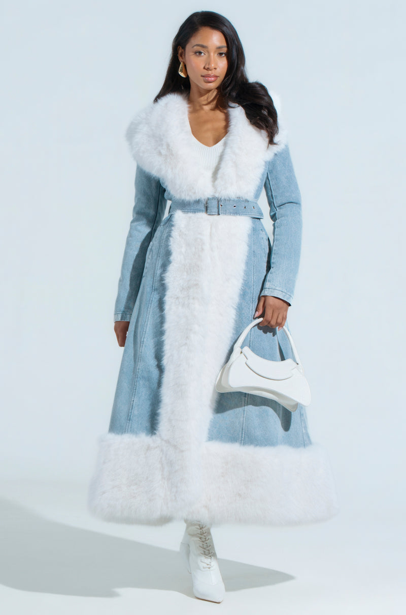 A woman confidently wears the VIVIAN FUR TRIM COAT IN DENIM, styled with an adjustable waist belt. She carries a white handbag, wears white heeled boots, and gold hoop earrings. Her long dark hair flows over her shoulders against a light, neutral background.
