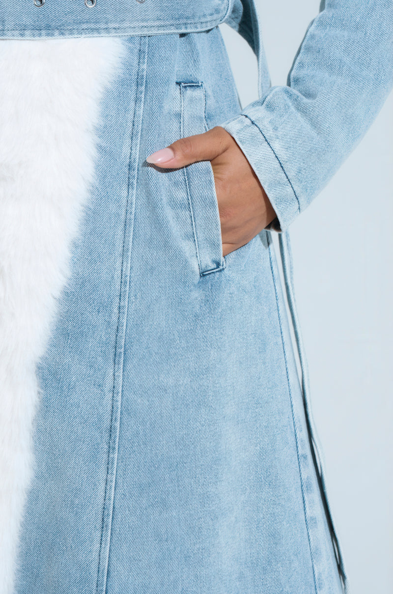 A person models the VIVIAN FUR TRIM COAT IN DENIM, featuring a light blue denim design and white faux fur trim along the side, with their hand in the pocket against a plain, light-colored background.