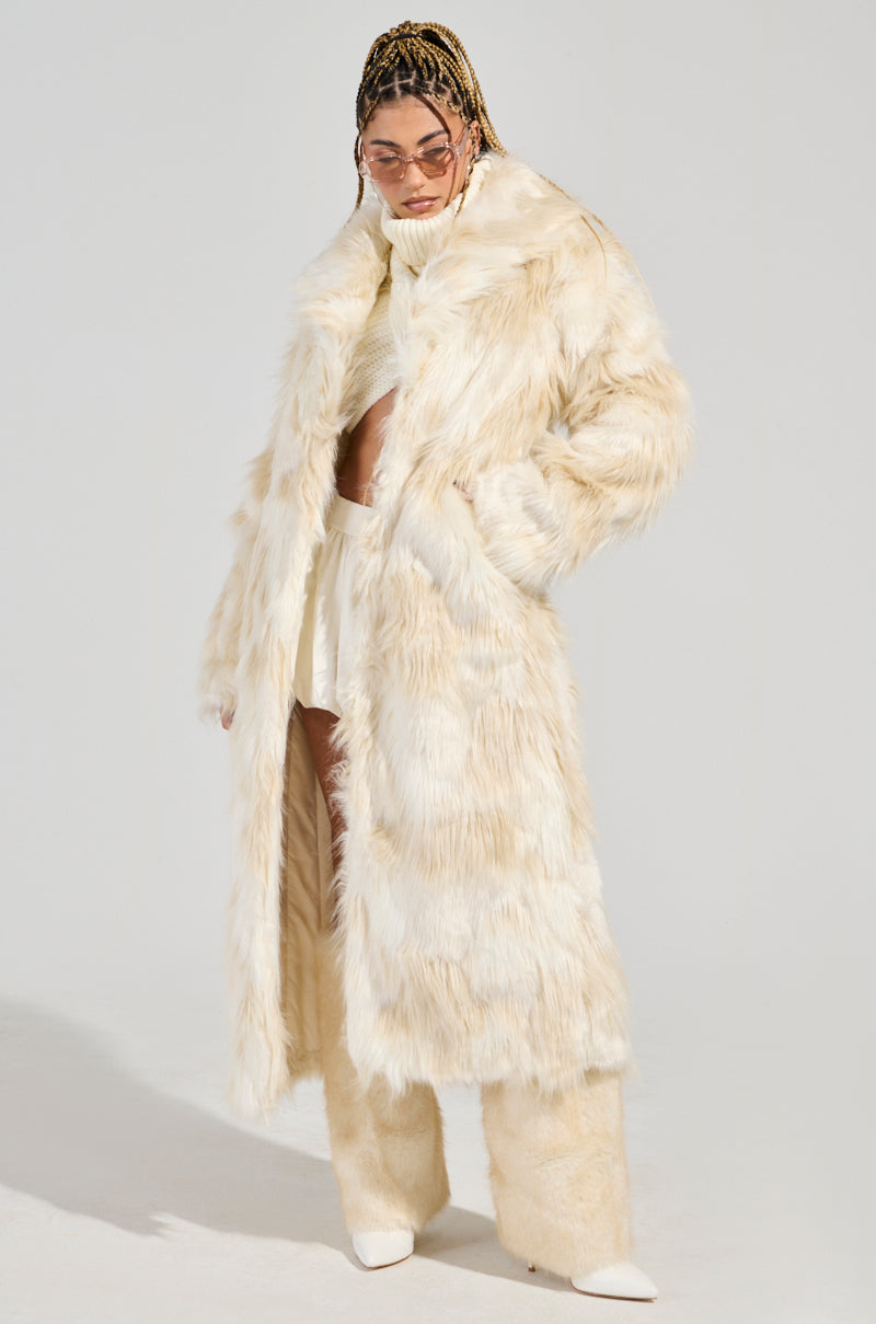 A person with braided hair and sunglasses is elegantly attired in the stylish BANKS FAUX FUR COAT, which showcases a longline silhouette and an oversized collar. The cream-colored ensemble, accompanied by coordinating pants and top, creates a fashionable contrast against a plain background.