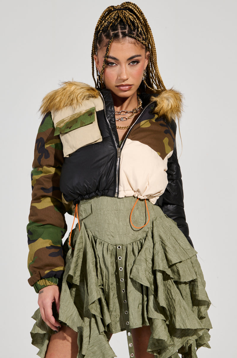 A person with braided hair is wearing the BENDITA CROPPED PATCHWORK PUFFER, which features a camouflage and color block pattern, faux fur trim, and a unique patchwork design. The green skirt is ruffled and layered, adorned with decorative buttons. The individual gazes confidently at the camera.