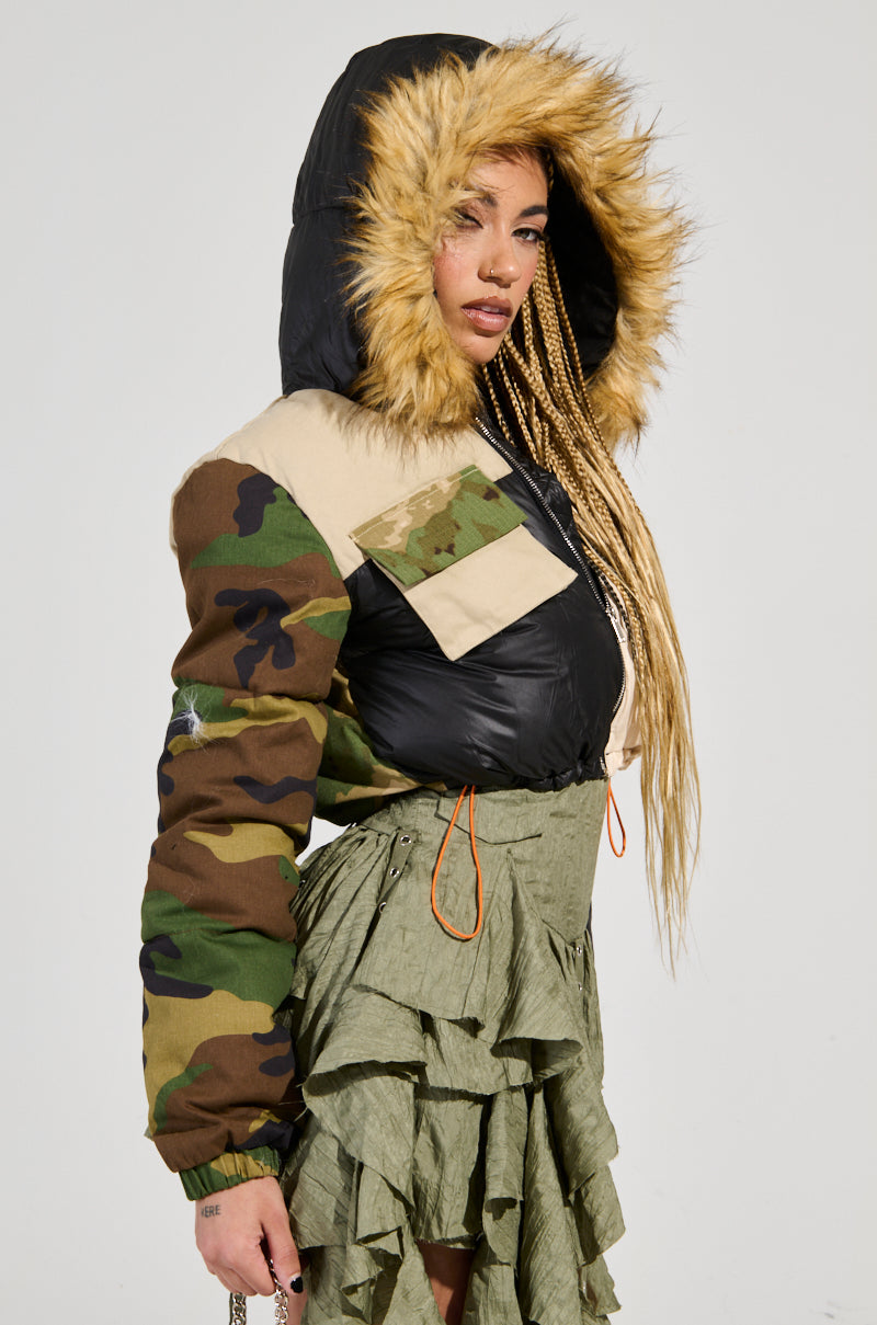 A person wearing the BENDITA CROPPED PATCHWORK PUFFER featuring a camo design and faux fur trim stands against a plain background, paired with a green ruffled skirt. Their long braided hair is visible, and they hold a small chain purse.