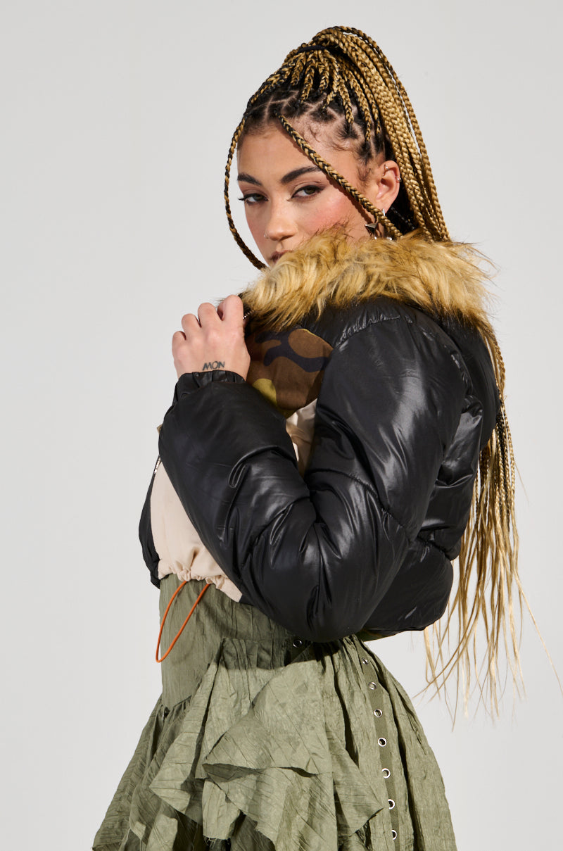A person with braided hair poses confidently, wearing the BENDITA CROPPED PATCHWORK PUFFER, a black jacket featuring a luxurious fur collar. Paired with a green, ruffled skirt, they gaze over their shoulder against a plain background, exuding elegant style and the subtle allure of faux fur trim.