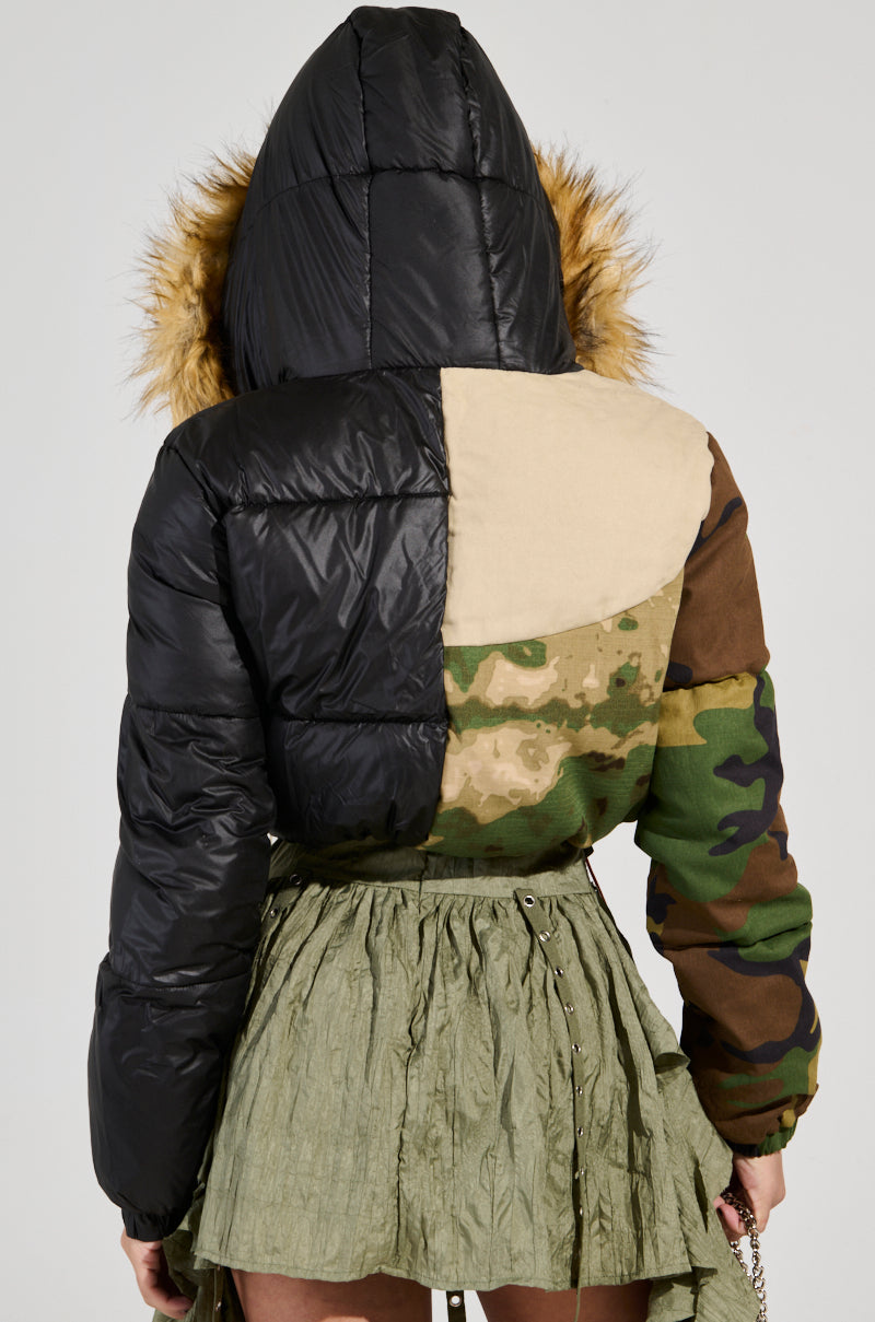 A person is wearing the BENDITA CROPPED PATCHWORK PUFFER jacket, featuring a faux fur trim hood and color block patterns in black, tan, and camouflage. They pair it with a green textured skirt while standing with their back to the camera.
