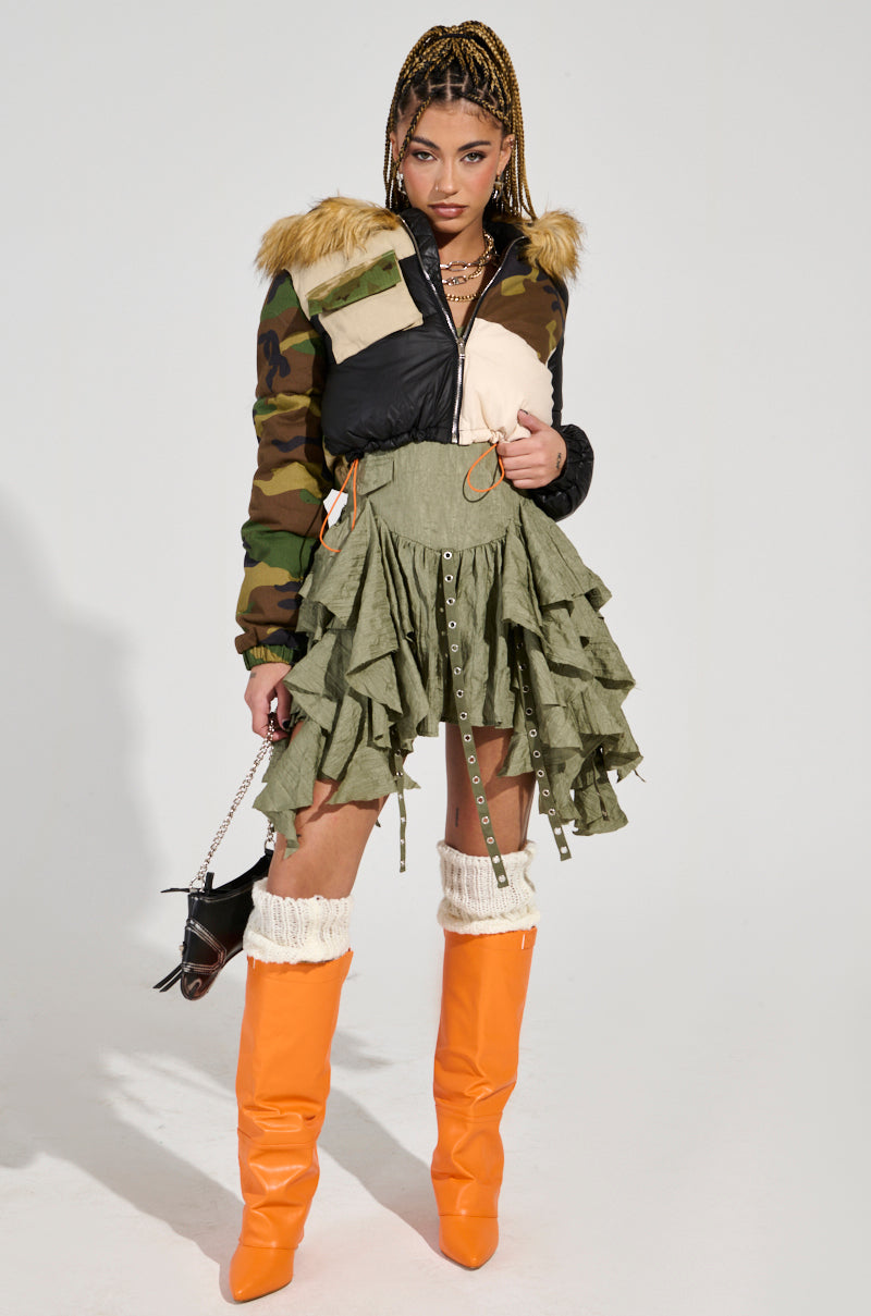 Wearing an outfit featuring the BENDITA CROPPED PATCHWORK PUFFER with a camouflage faux fur-trimmed design, an olive ruffled skirt, and vibrant orange knee-high boots, a person confidently poses while holding a small handbag. Their braided hair is adorned with gold chains and hoop earrings.