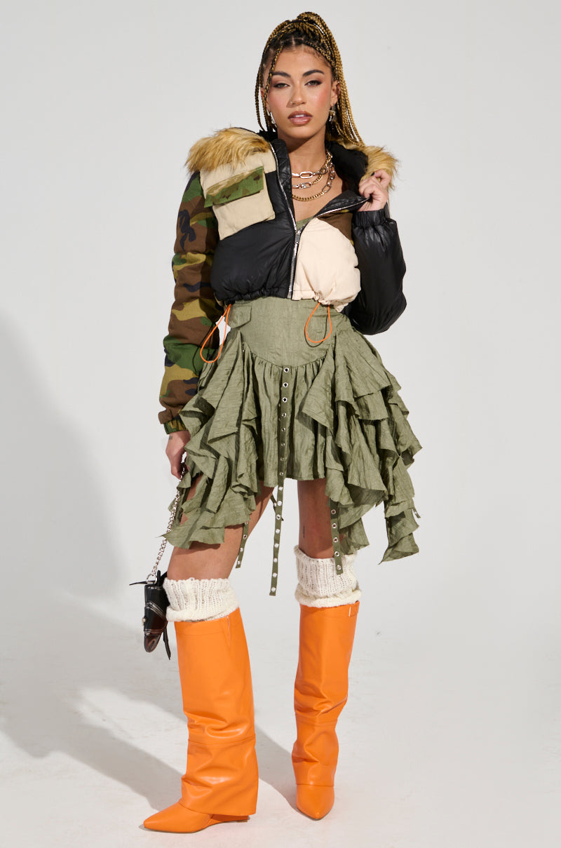 A person posing in a stylish outfit featuring the BENDITA CROPPED PATCHWORK PUFFER with faux fur trim, paired with a layered green skirt and bright orange knee-high boots. They have braided hair and wear multiple necklaces.
