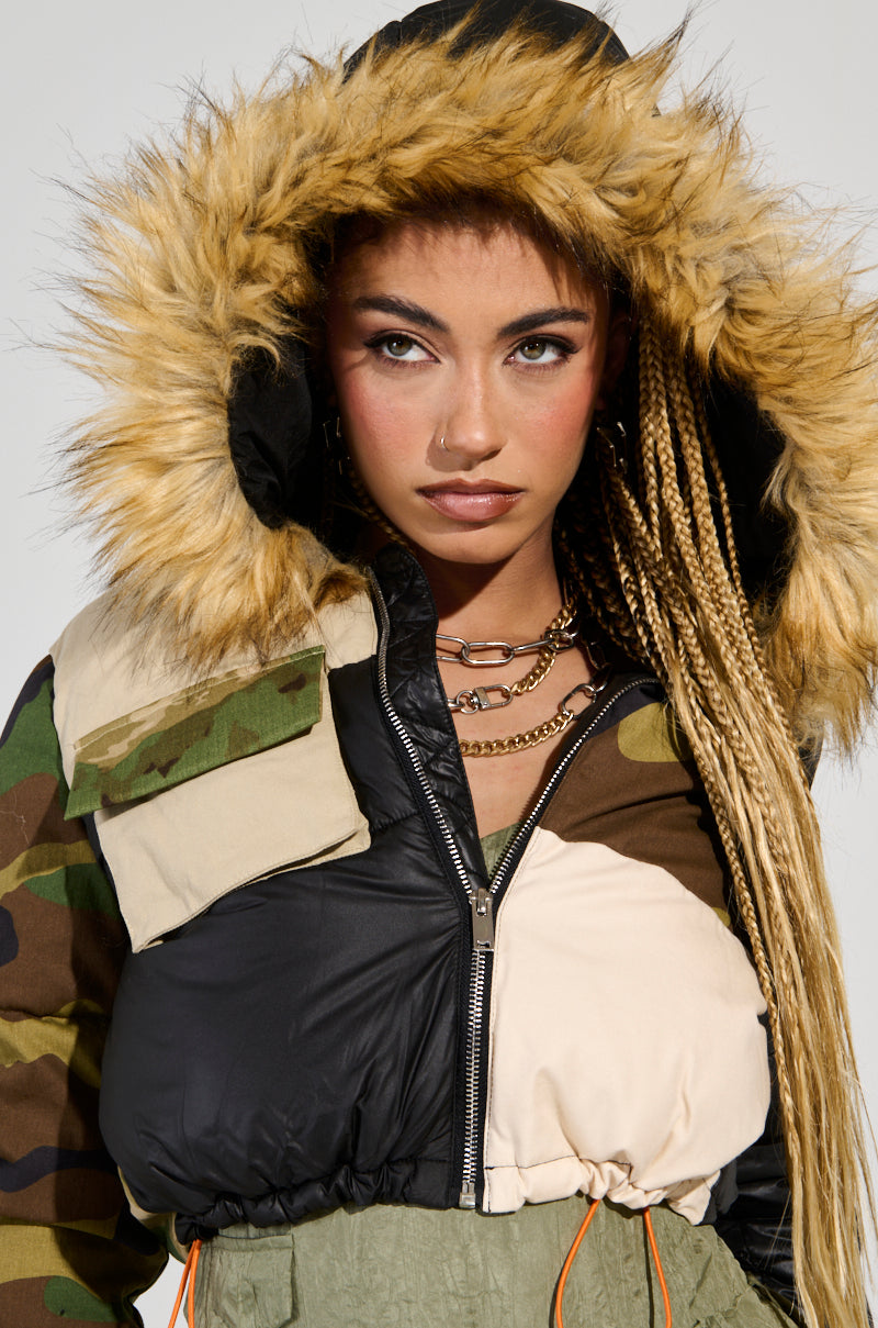 A young woman with long braided hair wears the BENDITA CROPPED PATCHWORK PUFFER, showcasing a camo and color block pattern with a faux fur trim on its large hood. Her expression is focused as she complements her look with layered gold chains. The jacket's earthy tones and varied textures add an edgy touch to her overall appearance.