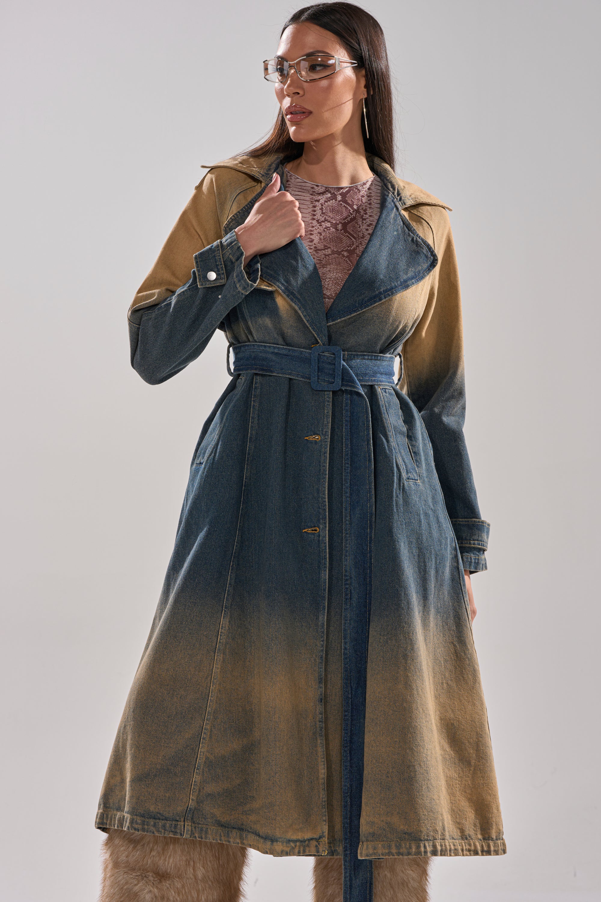 A woman wearing clear glasses and beige furry boots styles the ACID WASHED DENIM TRENCH over a textured mauve top, standing against a plain light background. 0