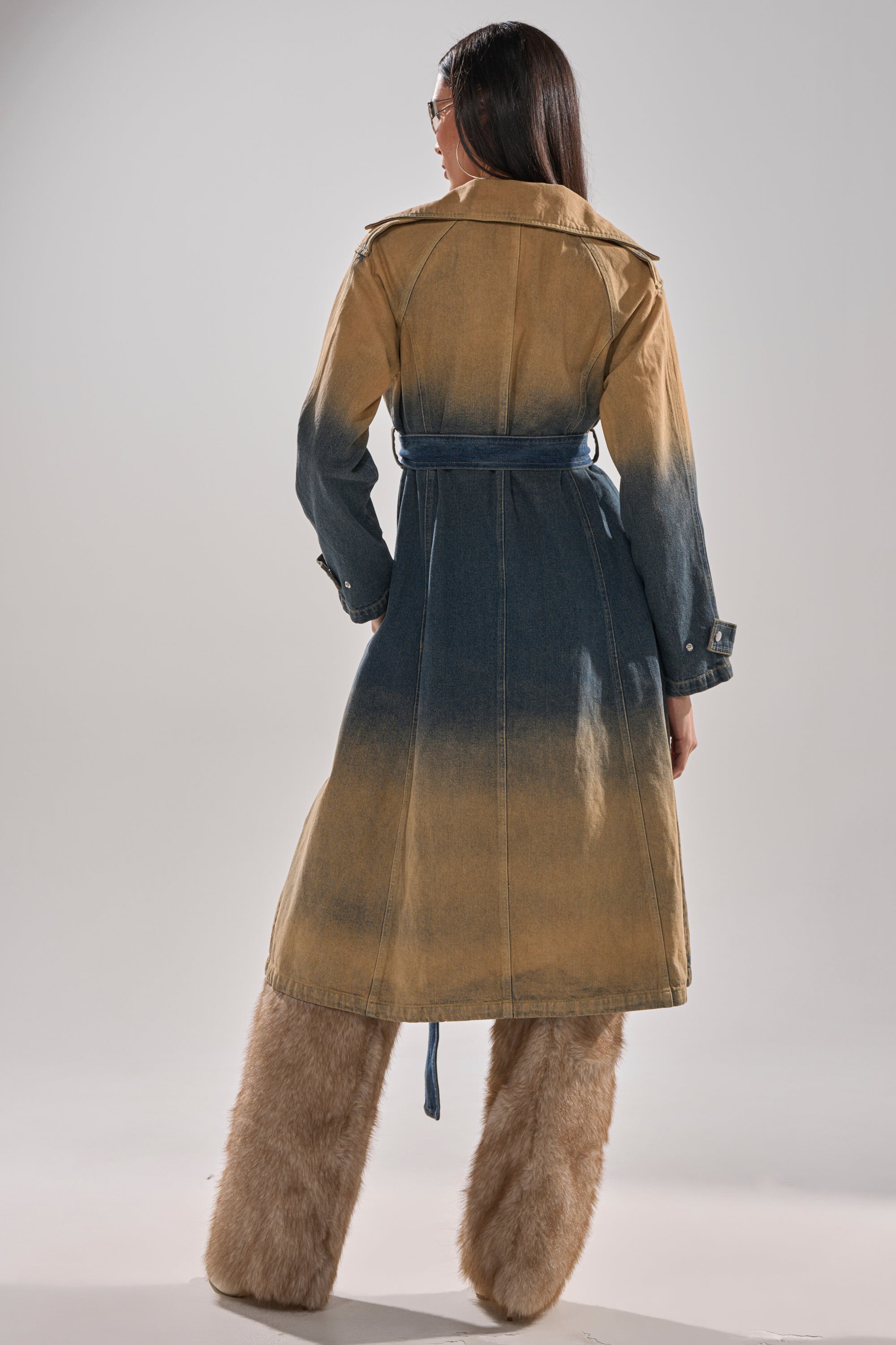 A person with long dark hair, seen from behind, wears the ACID WASHED DENIM TRENCH with a belted waist and oversized, fur-covered pants. The background is plain and light-colored. 9