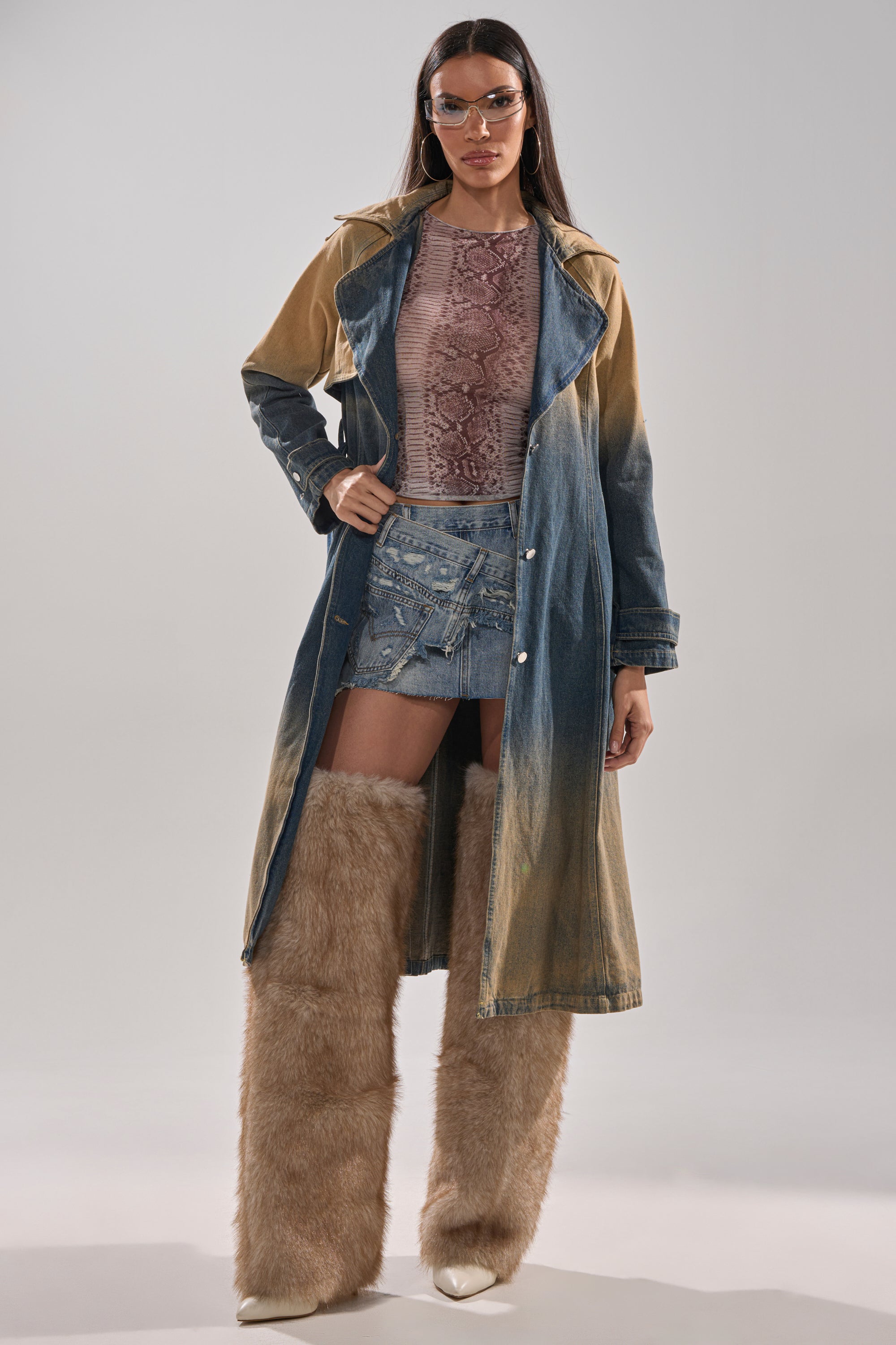 A woman in an ACID WASHED DENIM TRENCH, tinted glasses, a pink snakeskin-print top, a ripped denim miniskirt, and tan fur boots stands against a plain background. 11