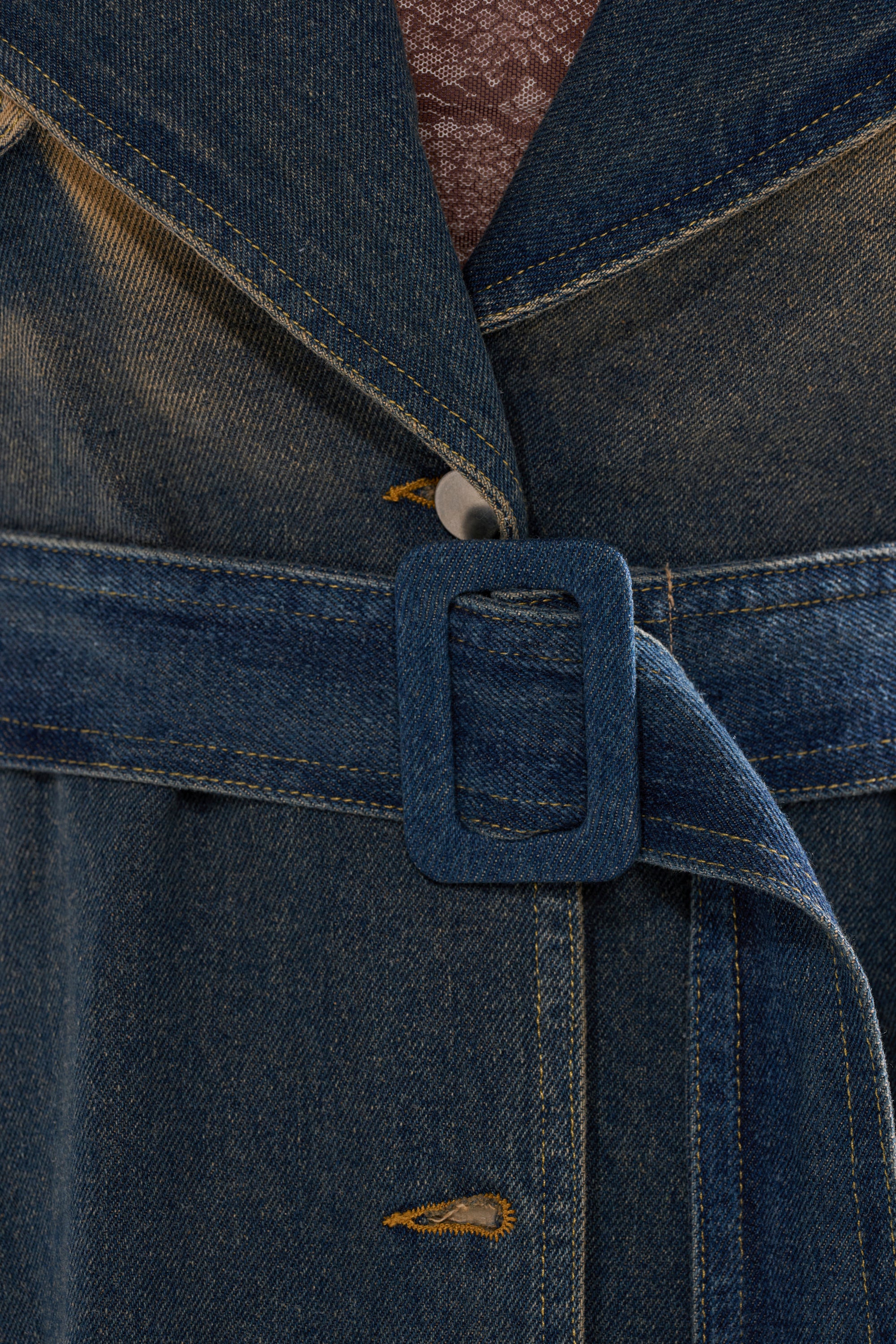Close-up of the ACID WASHED DENIM TRENCH featuring a matching denim belt with a rectangular buckle fastened through a metal buttonhole. A textured brownish fabric is visible underneath the coat. 12