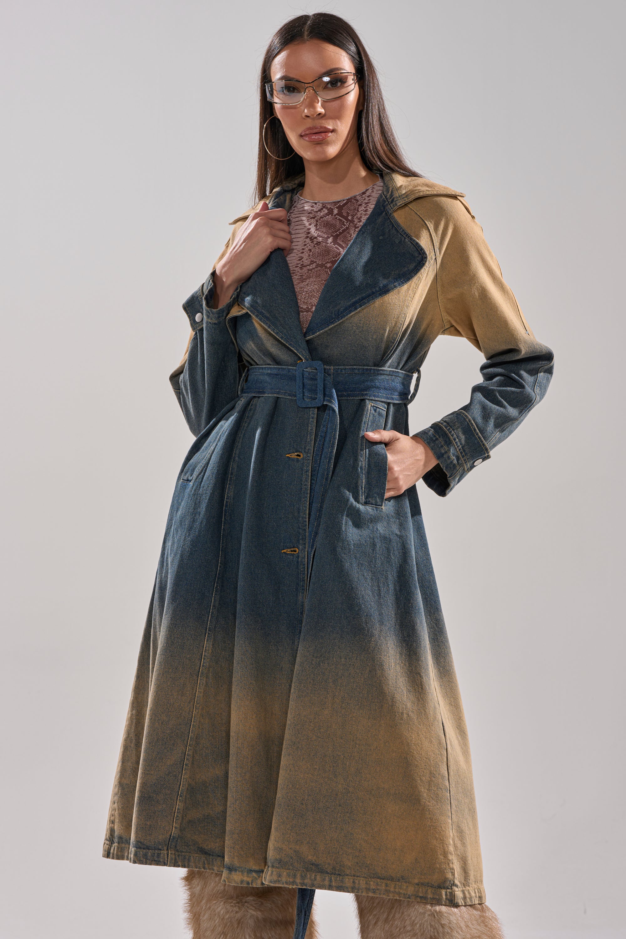 A woman in the ACID WASHED DENIM TRENCH with tan accents, clear visor sunglasses, large hoop earrings, a patterned top, and fur-trimmed boots stands against a plain light background. 1