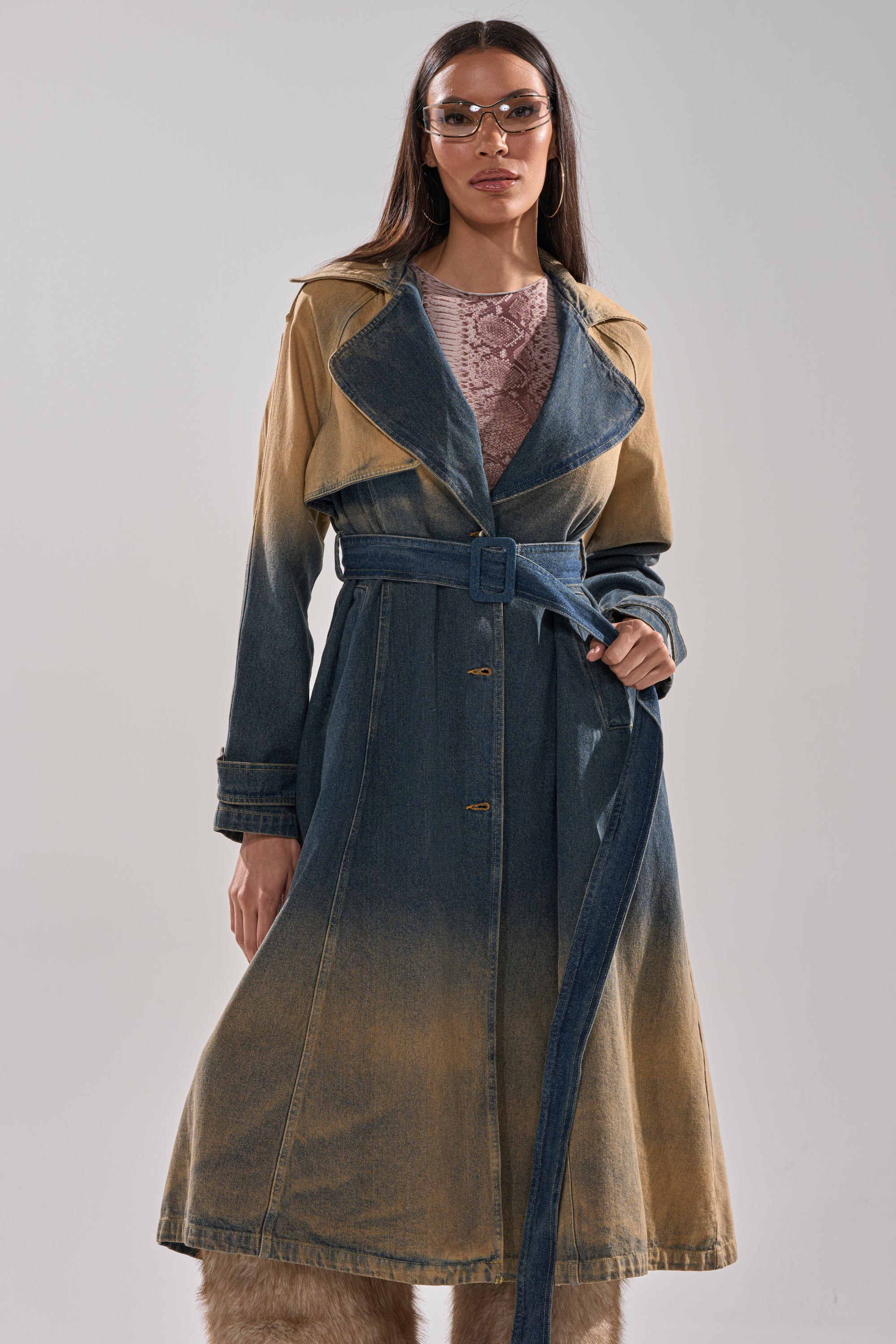 A woman with glasses wears the ACID WASHED DENIM TRENCH, featuring a belted waist and a faded gradient wash. She poses confidently with long hair and a serious expression against a plain background. 2