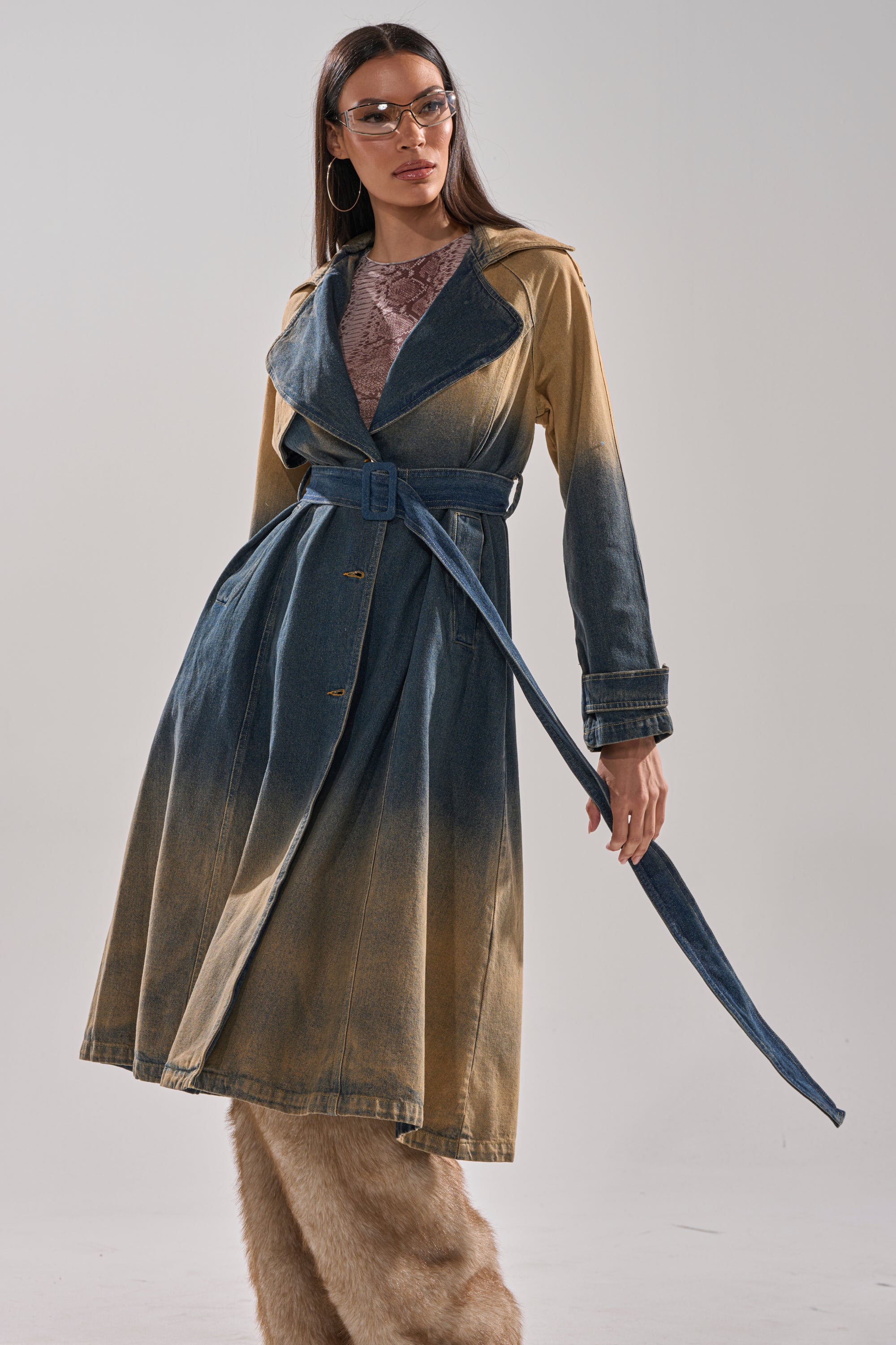 A woman confidently poses against a plain gray background, wearing clear glasses and the ACID WASHED DENIM TRENCH over a lace top, paired with wide, furry tan pants. 3