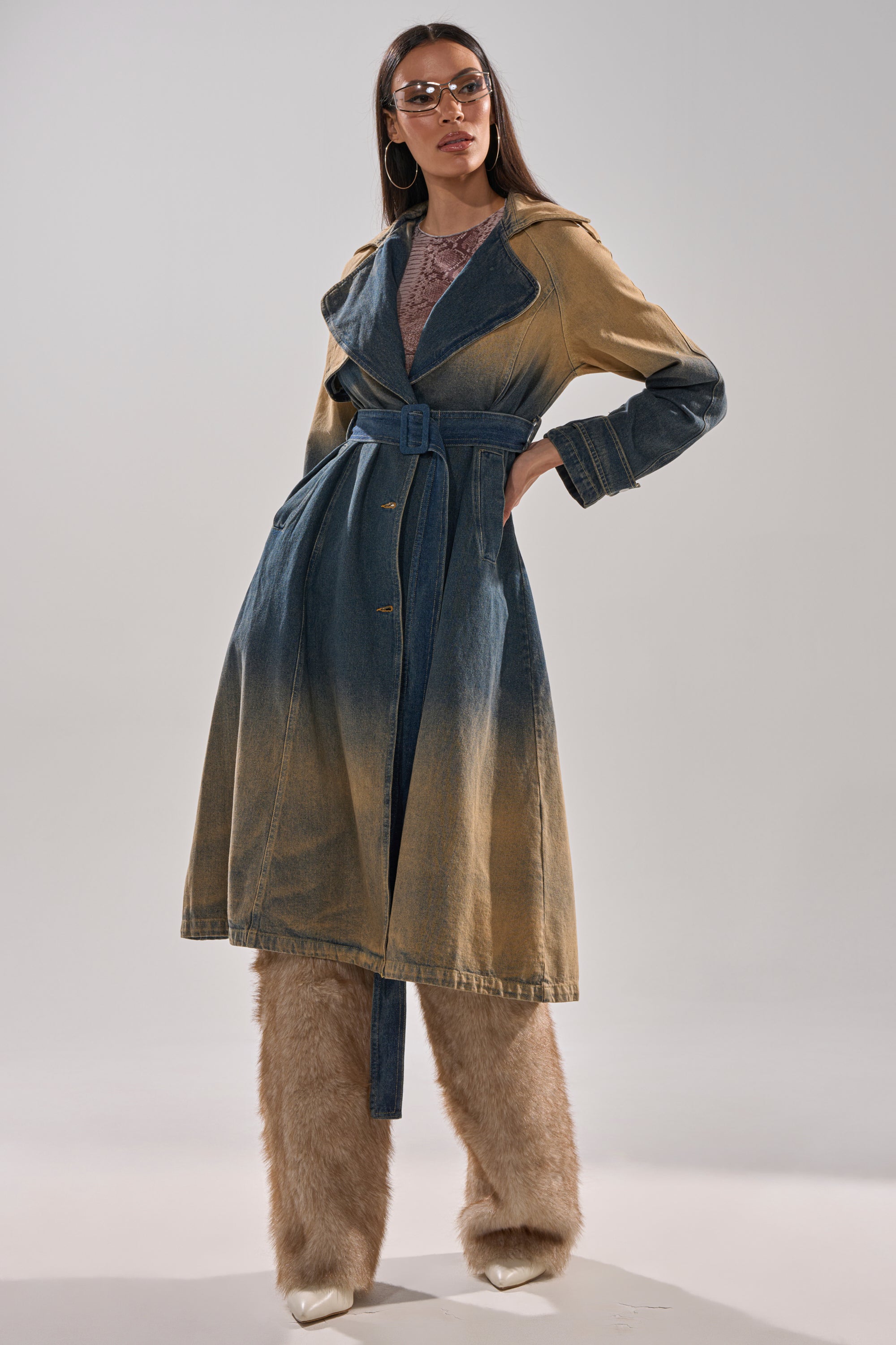 A woman poses confidently in the ACID WASHED DENIM TRENCH with wide lapels, large sunglasses, and tan furry pants over white pointed shoes, set against a plain, light-colored background. 4
