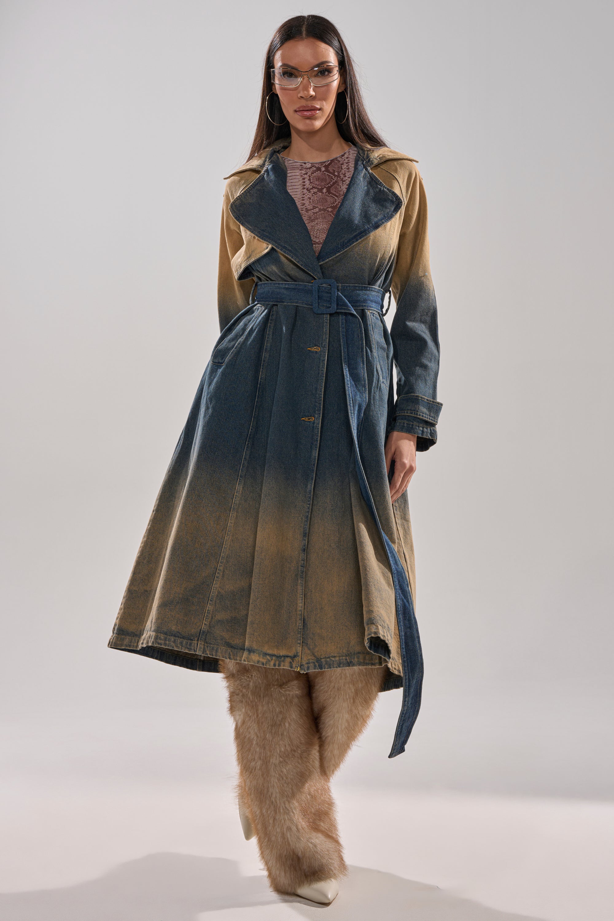 A woman poses in the ACID WASHED DENIM TRENCH, belted over a mauve top, beige furry pants, white shoes, and clear glasses, set against a plain light background. 5