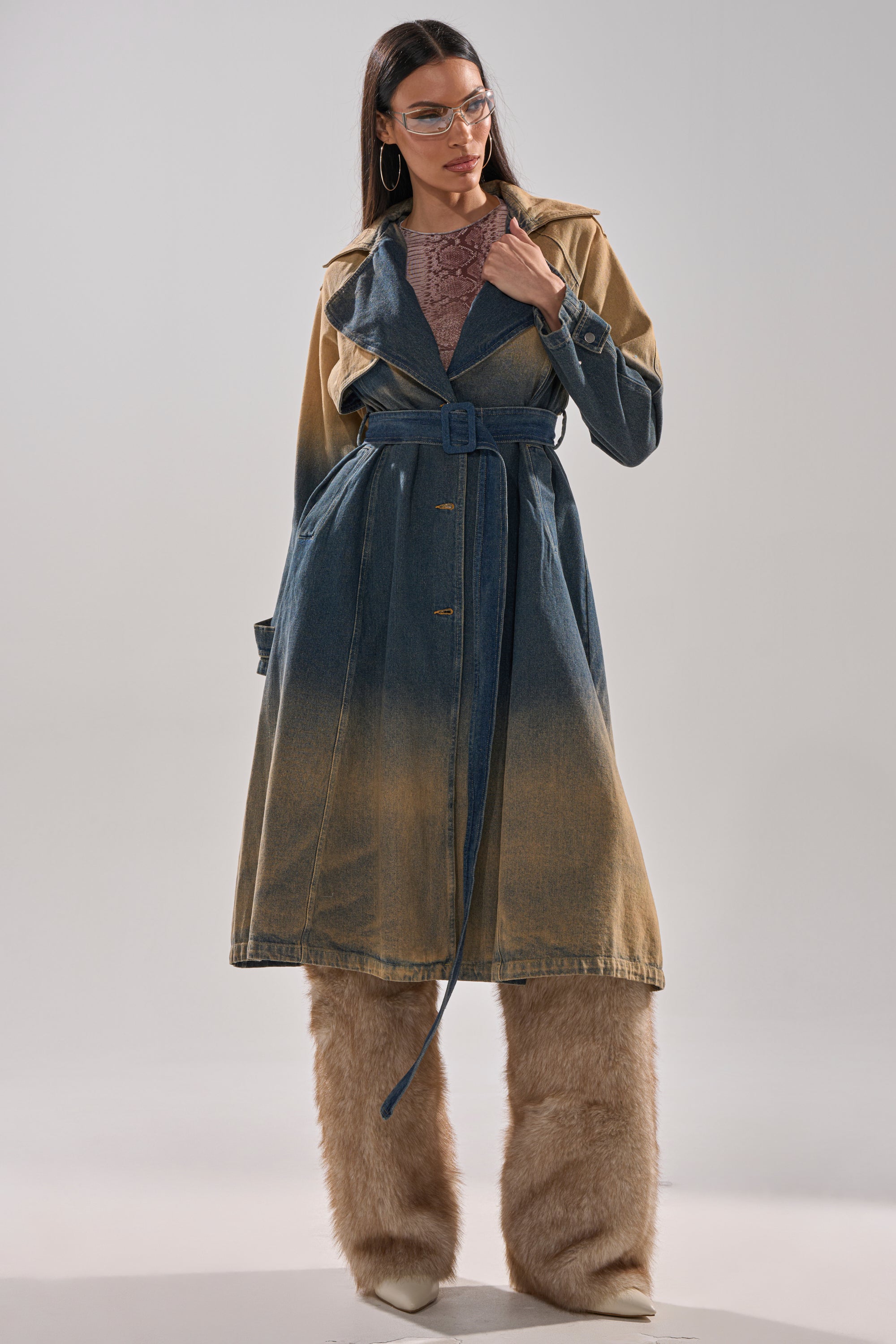 A woman poses confidently against a plain background wearing the ACID WASHED DENIM TRENCH, furry tan pants, white pointed shoes, large hoop earrings, clear glasses, and a sheer patterned top. 6