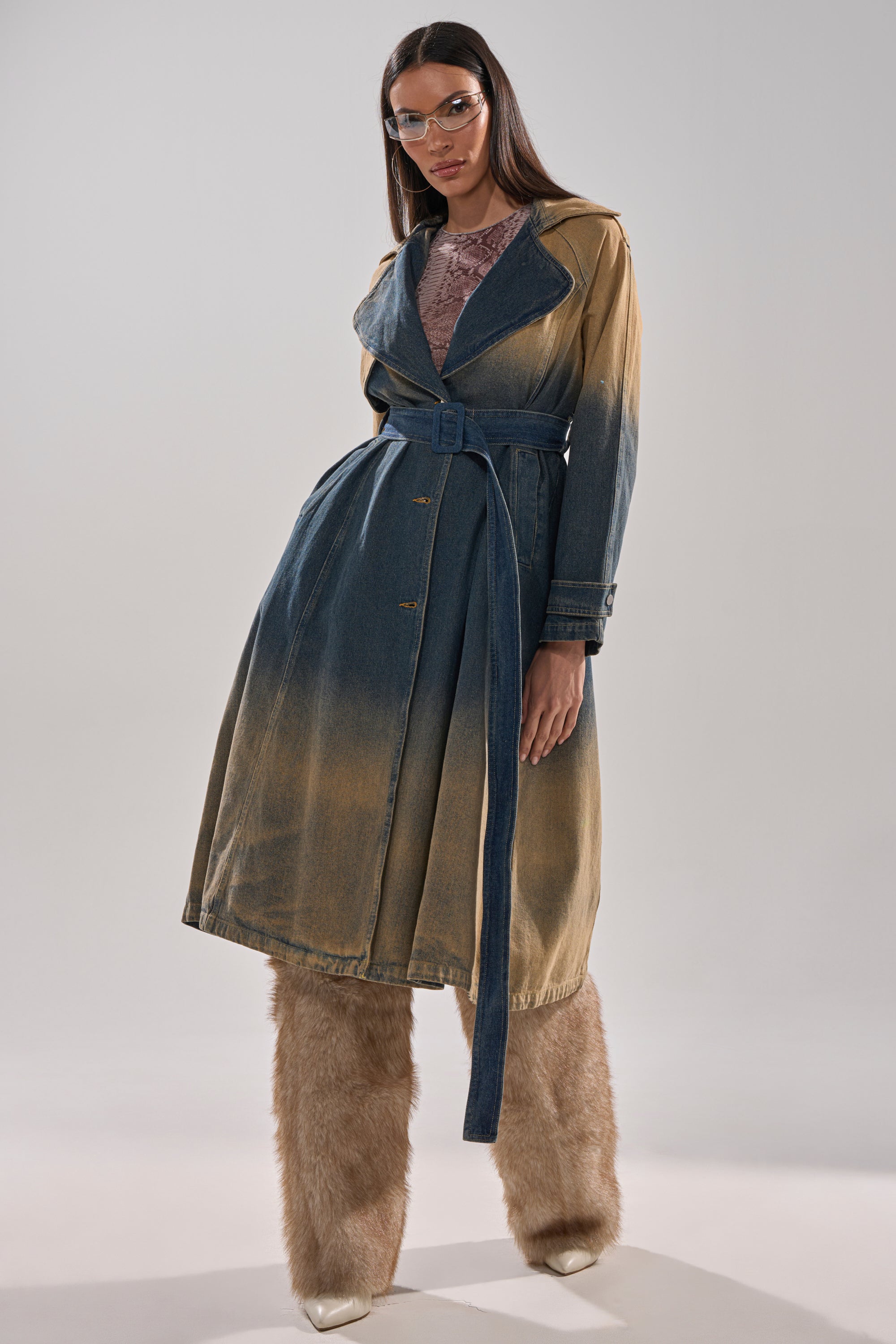 A person in the ACID WASHED DENIM TRENCH, belted at the waist, pairs it with light brown furry pants, pointed white shoes, and clear glasses, standing against a plain light background. 7