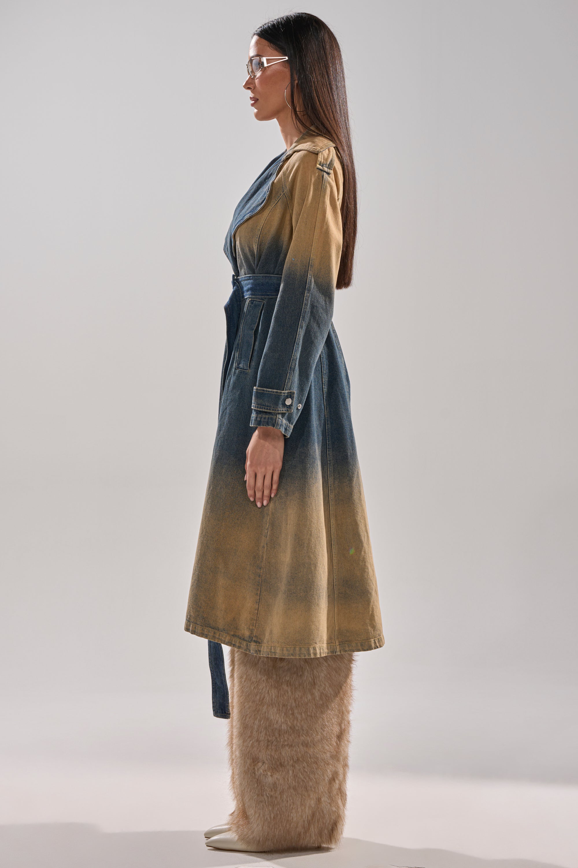 A woman stands in profile, confidently wearing the ACID WASHED DENIM TRENCH with a tie belt, brown faux-fur pants, white pointed shoes, and clear glasses. Her straight hair is loose as she poses against a plain background. 8