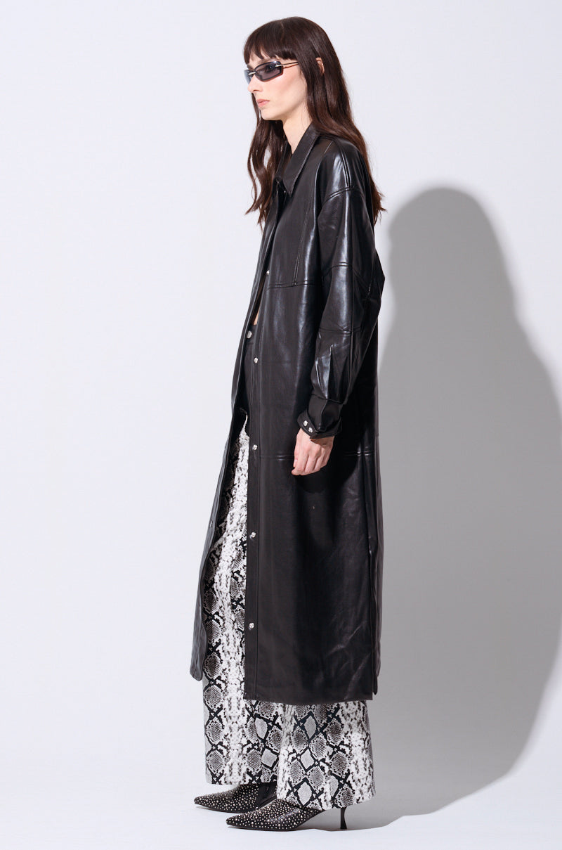 A woman with long dark hair wears dark sunglasses, the OSAKA BOMBER TRENCH in a maxi length silhouette, and snakeskin-patterned pants. She stands in a side profile against a plain background, her right hand resting by her side.