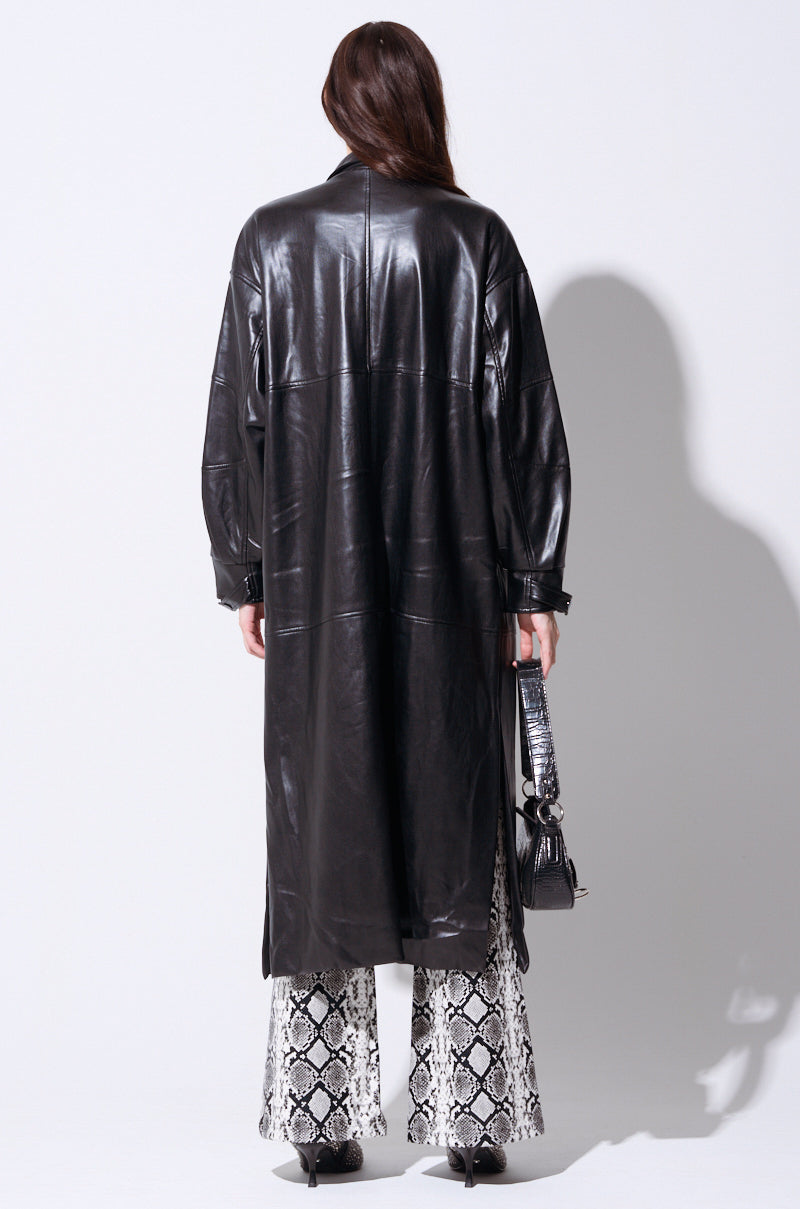 A person with long brown hair is shown from the back, wearing an OSAKA BOMBER TRENCH in a maxi-length silhouette made of black leather over black and white snake print flared pants. They are holding a small black handbag in their right hand and standing against a plain white background.