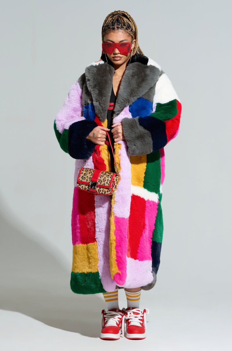 A person wearing the STUDIO 24 FAUX FUR COAT in an oversized silhouette showcases a vibrant patchwork design, complemented by red sunglasses and a small patterned handbag. Their hair is styled in braids, and they are sporting red and white sneakers paired with striped socks, all set against a plain background.