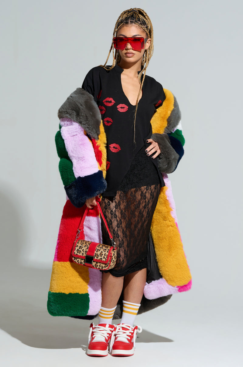 A fashionable person stands confidently in the STUDIO 24 FAUX FUR COAT, featuring a color-block pattern, their black outfit adorned with red lips designs. Red sunglasses and sneakers complete the look, while a small leopard print bag and braided hair add flair to their oversized silhouette.