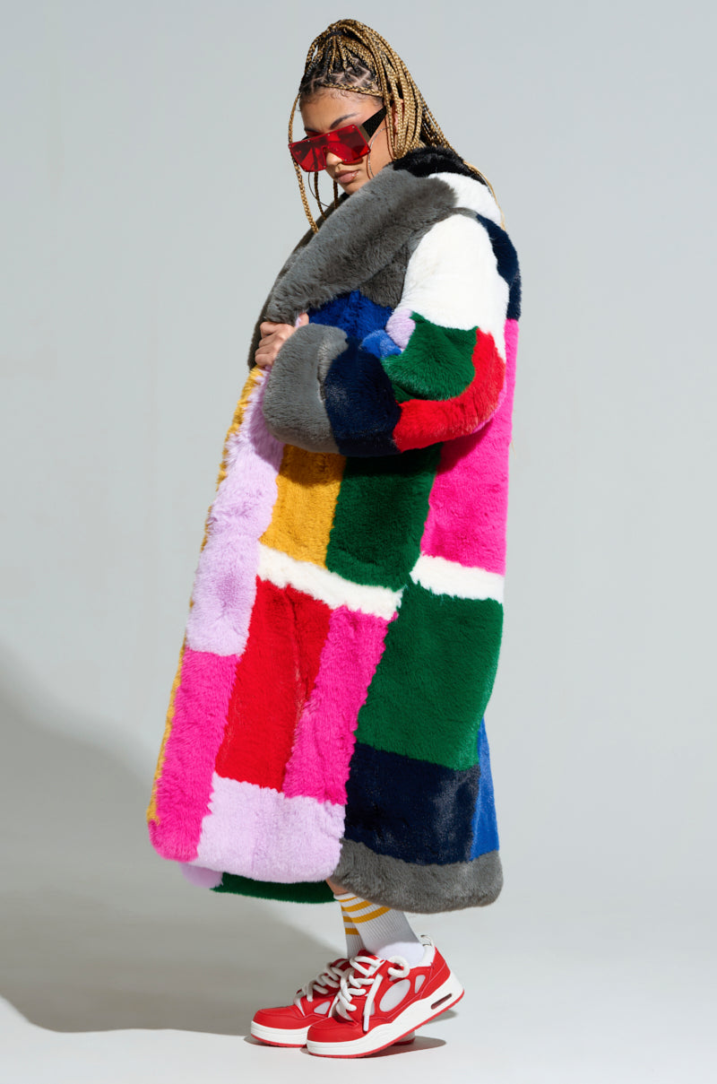 A person wearing the STUDIO 24 FAUX FUR COAT in an oversized multicolored patchwork style stands in profile. They have braided hair and wear red sunglasses, yellow-striped socks, and red sneakers against a plain gray background.