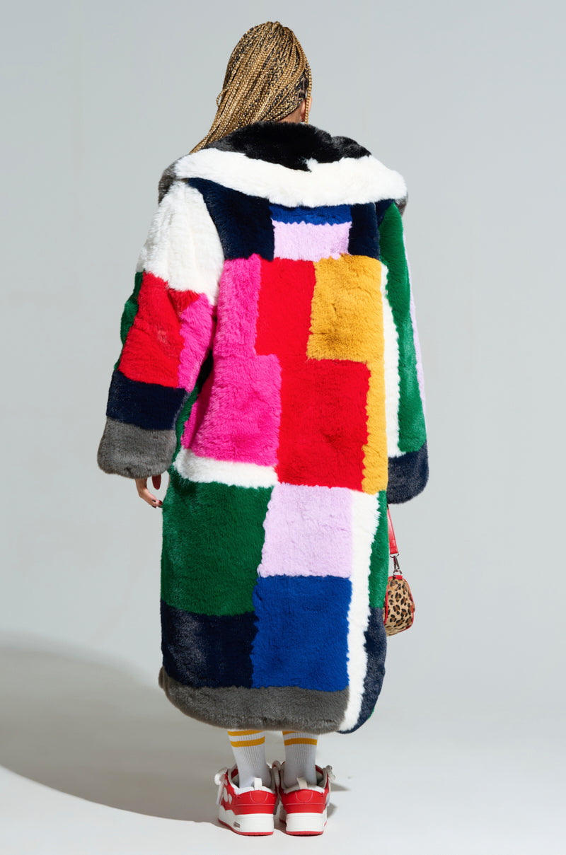 A person with braided hair stands facing away, showcasing the STUDIO 24 FAUX FUR COAT with a bold color-block pattern in pink, green, blue, red, and white. They accessorize with a leopard print bag and complement their look with yellow socks paired with red and white sneakers.