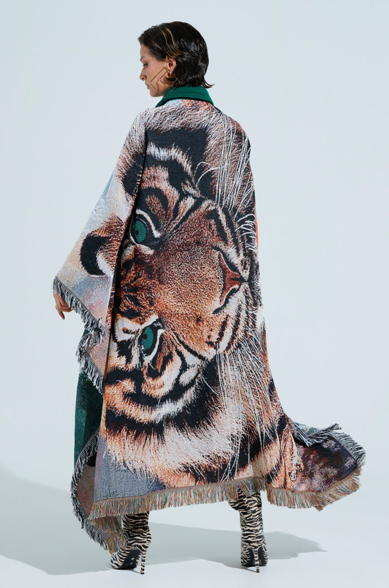 Wearing a large, fringed shawl with a vivid tiger face design, the person drapes it over their shoulders alongside zebra-patterned shoes. They complement this look with an evergreen wool-blend bomber jacket named "FIRST ROUND DRAFT PICK TAPESTRY BOMBER" against a simple, light background.