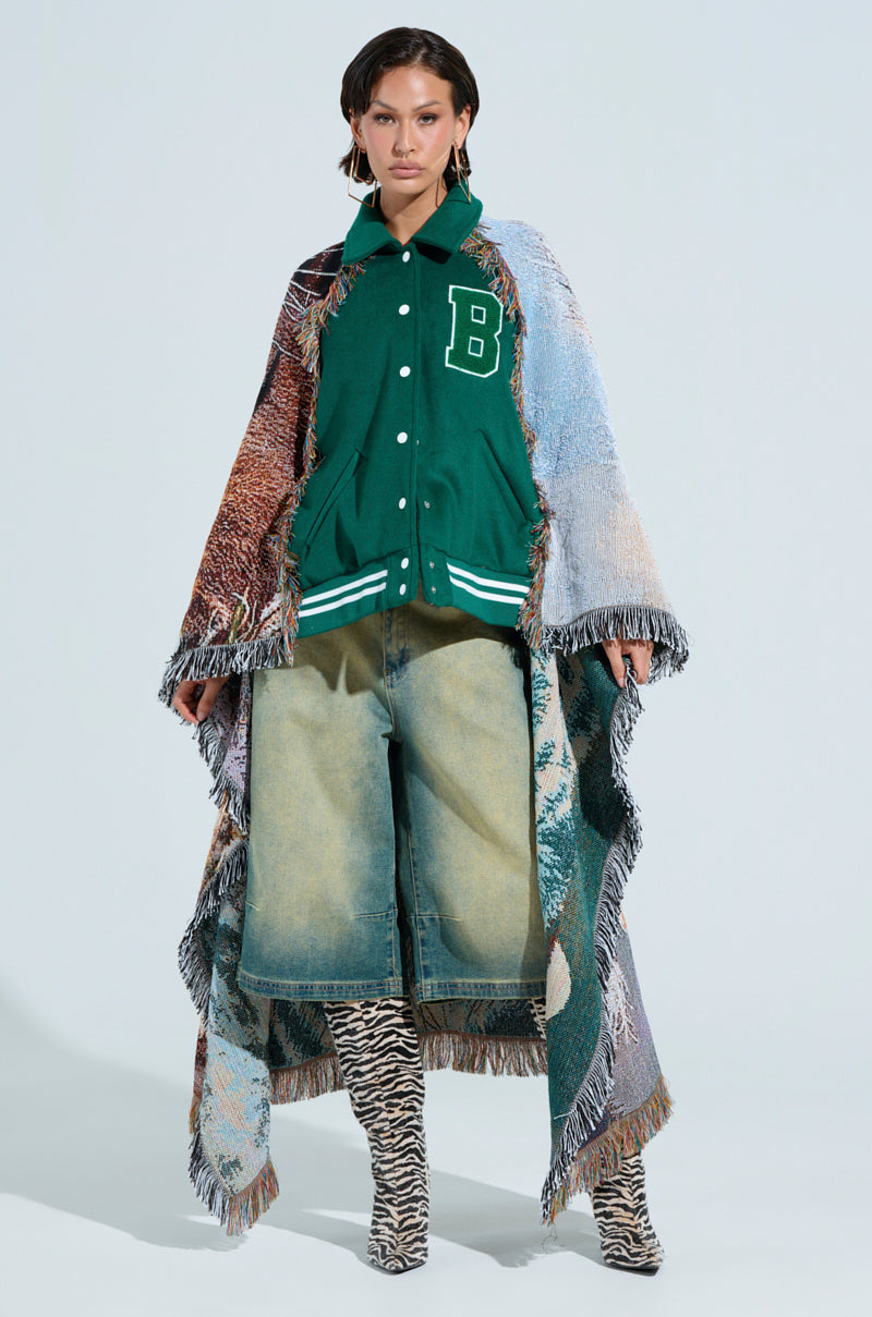 A person confidently wears the FIRST ROUND DRAFT PICK TAPESTRY BOMBER, oversized denim shorts, zebra-striped boots, and a fringed multicolored blanket draped around them. The plain light background highlights their eclectic fashion style.